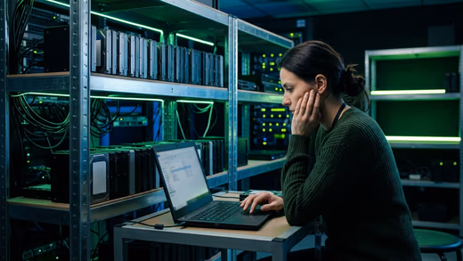 Une technicienne en pull vert vérifie une sauvegarde sur un ordinateur portable, devant des étagères de disques durs