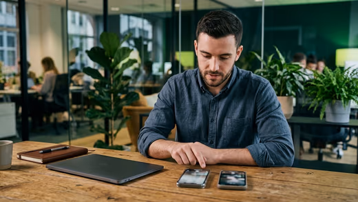 Jeune responsable acquisition comparant deux smartphones sur un bureau en bois, dans un open space aux accents verts